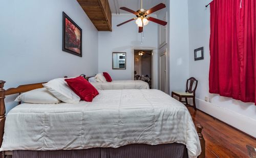 Cozy bedroom with two beds, red accents, a ceiling fan, and a window with red curtains. Warm wooden floors.