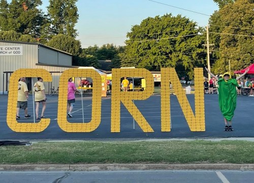 A person in a corn costume stands next to large letters spelling "CORN" at an outdoor event with people in the background.