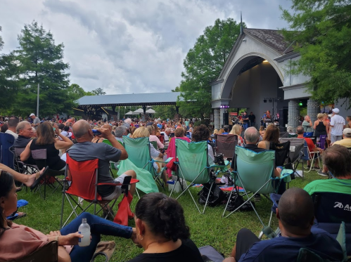 A large crowd sits on lawn chairs at an outdoor concert, with a stage and trees in the background under a cloudy sky.