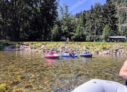 Three people in inflatable tubes float on a clear river, surrounded by trees and a rocky shoreline.