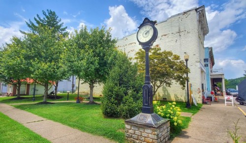 A vintage clock stands in a green park, surrounded by trees and a historic building under a blue sky with clouds.