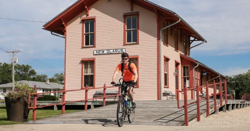 A cyclist in an orange shirt rides past a historic train station labeled "New Glarus" on a sunny day.