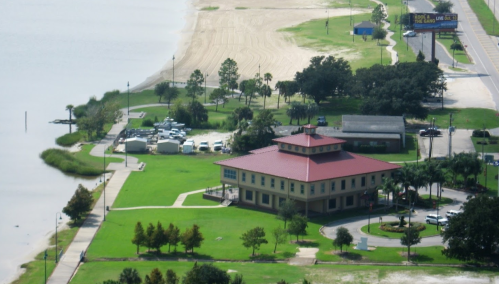 Aerial view of a park with a red-roofed building, green lawns, and a sandy beach by a lake.