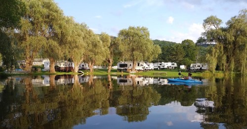 A serene campsite by a lake, featuring RVs and kayakers surrounded by lush trees and reflections on the water.