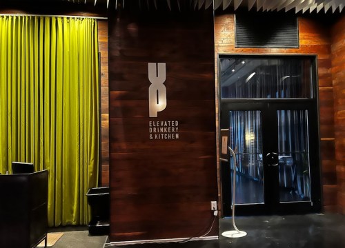 Interior of a restaurant with a wooden wall, green curtains, and a sign reading "Elevated Drinkery & Kitchen."