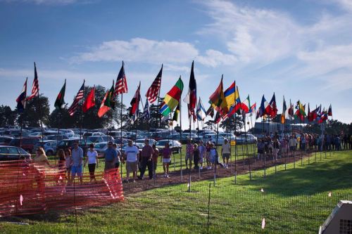 A line of people walking past a row of flags from various countries under a clear blue sky.