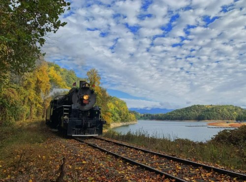 A vintage steam train travels along a scenic lakeside track, surrounded by autumn foliage and a cloudy sky.
