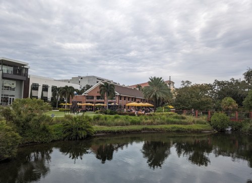 A scenic view of a restaurant by a pond, surrounded by greenery and palm trees, under a cloudy sky.