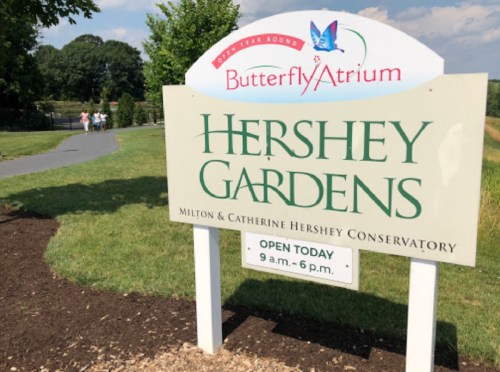 Sign for Hershey Gardens featuring the Butterfly Atrium, with visitors walking in the background on a sunny day.