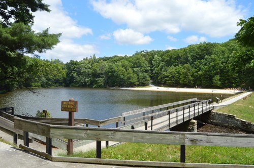 A serene lake surrounded by trees, with a wooden bridge leading to a sandy beach under a partly cloudy sky.