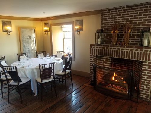 Cozy dining room with a round table set for dinner, surrounded by chairs and a brick fireplace with a warm fire.