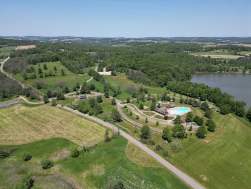 Aerial view of a green landscape with a pool, trees, and a lake, surrounded by rolling hills under a clear blue sky.