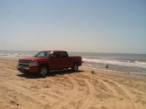 A red pickup truck parked on a sandy beach with ocean waves in the background and a person playing in the water.