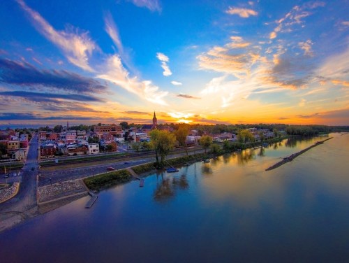 A vibrant sunset over a riverside town, with colorful clouds reflecting on the water and buildings lining the shore.