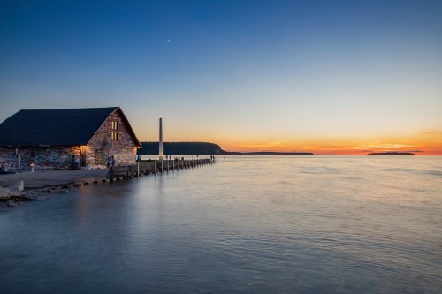 A serene sunset over calm waters, featuring a rustic stone building and a distant shoreline.