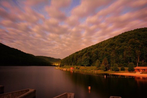 A serene lake surrounded by lush green hills under a cloudy night sky, reflecting soft light on the water's surface.