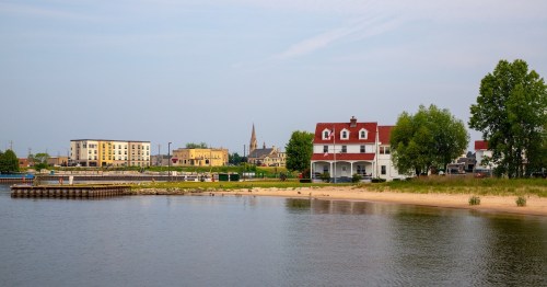 A serene waterfront scene featuring a sandy beach, a white house, and buildings in the background under a clear sky.