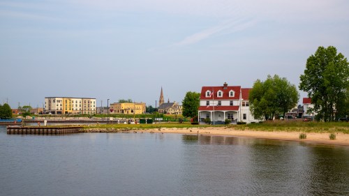 A serene lakeside view featuring a white house, sandy beach, and distant buildings under a clear sky.