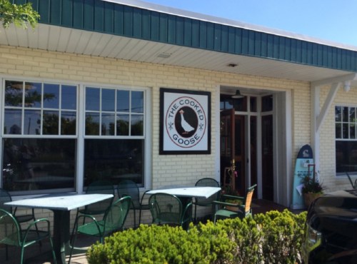 Exterior of "The Cooked Goose" restaurant with a sign, green chairs, and a sunny day.