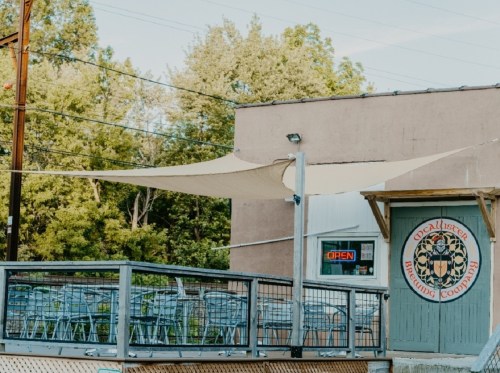 A brewery with outdoor seating under a shade sail, surrounded by trees, and an "Open" sign in the window.