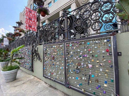 A decorative fence adorned with numerous colorful locks and hanging plants, set against a building backdrop.