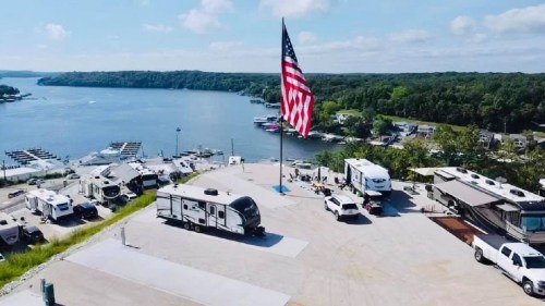 Aerial view of an RV park by a lake, featuring a large American flag and several parked RVs.