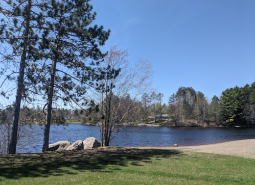 A serene lakeside view with trees, rocks, and clear blue skies, surrounded by lush greenery.