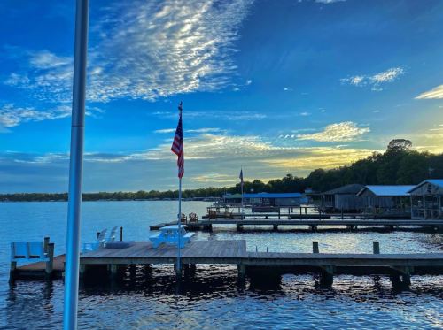A serene lakeside view at sunset, featuring docks, an American flag, and a colorful sky with scattered clouds.