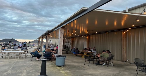 Outdoor seating area with string lights, people dining, and a cloudy sky in the background.