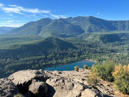 A scenic view of mountains and a lake surrounded by lush green forests under a clear blue sky.