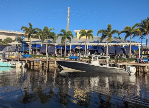 A boat glides past a waterfront restaurant with palm trees and outdoor seating under blue skies.