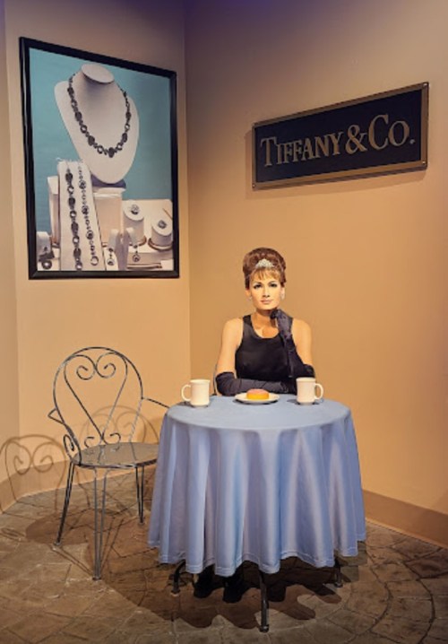 A mannequin resembling a woman sits at a table with coffee and a pastry, next to a Tiffany & Co. display.