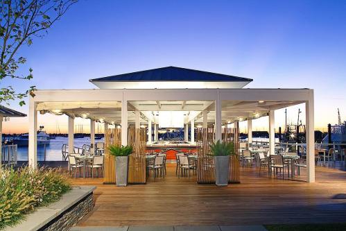 A waterfront restaurant with outdoor seating, illuminated under a twilight sky, surrounded by boats and water.