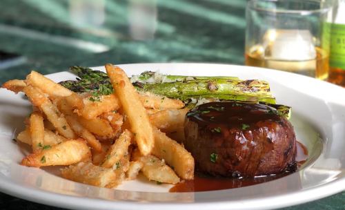 A plate featuring a grilled steak with sauce, asparagus, and crispy fries, accompanied by a glass of whiskey.