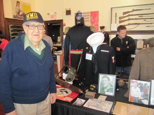 An elderly veteran stands beside a display of military uniforms and artifacts in a museum setting.