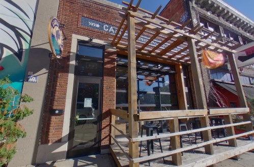 Exterior of a brick building with a wooden patio and large glass doors, featuring colorful signage above.