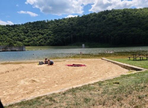 A sandy beach by a calm lake, with people relaxing and swimming, surrounded by green hills under a blue sky.