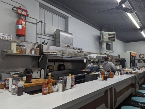 A diner kitchen with a grill, stacked plates, condiments, and a cook preparing food at the counter.
