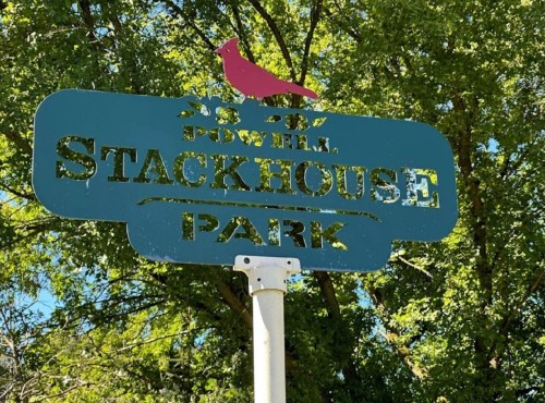 Sign for Stackhouse Park with a red bird design, surrounded by green trees.