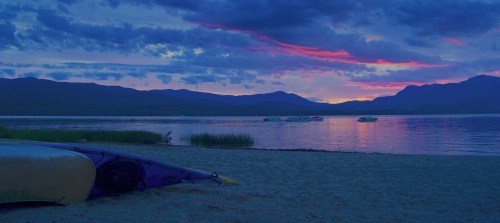 A serene lakeside scene at dusk, with a kayak on the sandy shore and colorful clouds reflecting on the water.
