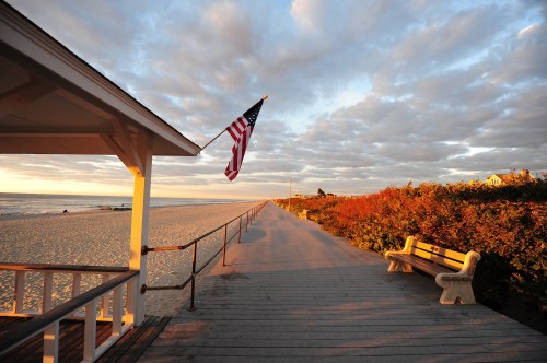 A beachside walkway at sunset, featuring a flag, benches, and vibrant foliage along the path.