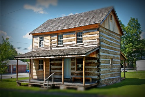A rustic two-story log cabin with a porch, surrounded by green grass and trees under a clear blue sky.