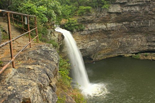 A waterfall cascades into a serene pool, surrounded by rocky cliffs and lush greenery. A path runs alongside the edge.