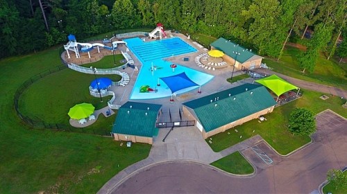 Aerial view of a swimming pool area with slides, lounge chairs, and colorful umbrellas surrounded by greenery.