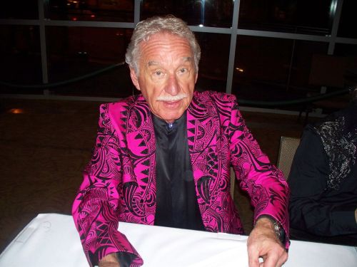 A man in a vibrant pink patterned jacket sits at a table, smiling and looking at the camera.