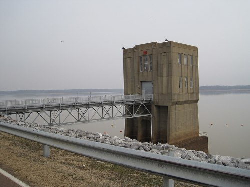 A concrete water control tower stands over a calm lake, with a walkway and rocky shoreline in the foreground.