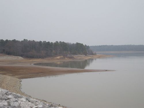 A calm lake with a sandy shore, surrounded by trees, under a cloudy sky. The water level appears low.