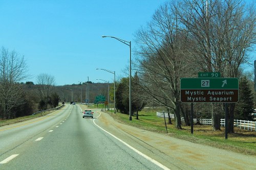 Road sign indicating exit 90 for Mystic Aquarium and Mystic Seaport, with a clear blue sky and trees lining the road.