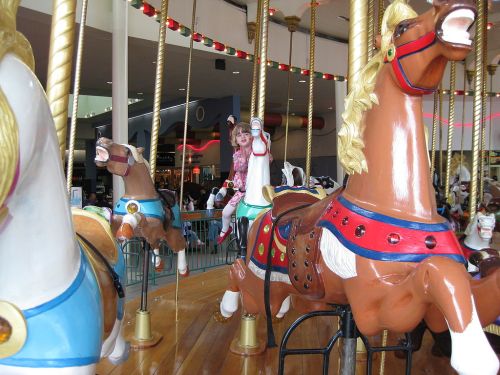 A child rides a colorful carousel horse, surrounded by other painted horses in a lively amusement setting.