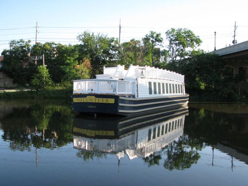 A boat named "Volunteer" docked on calm water, reflecting its image and surrounded by greenery.